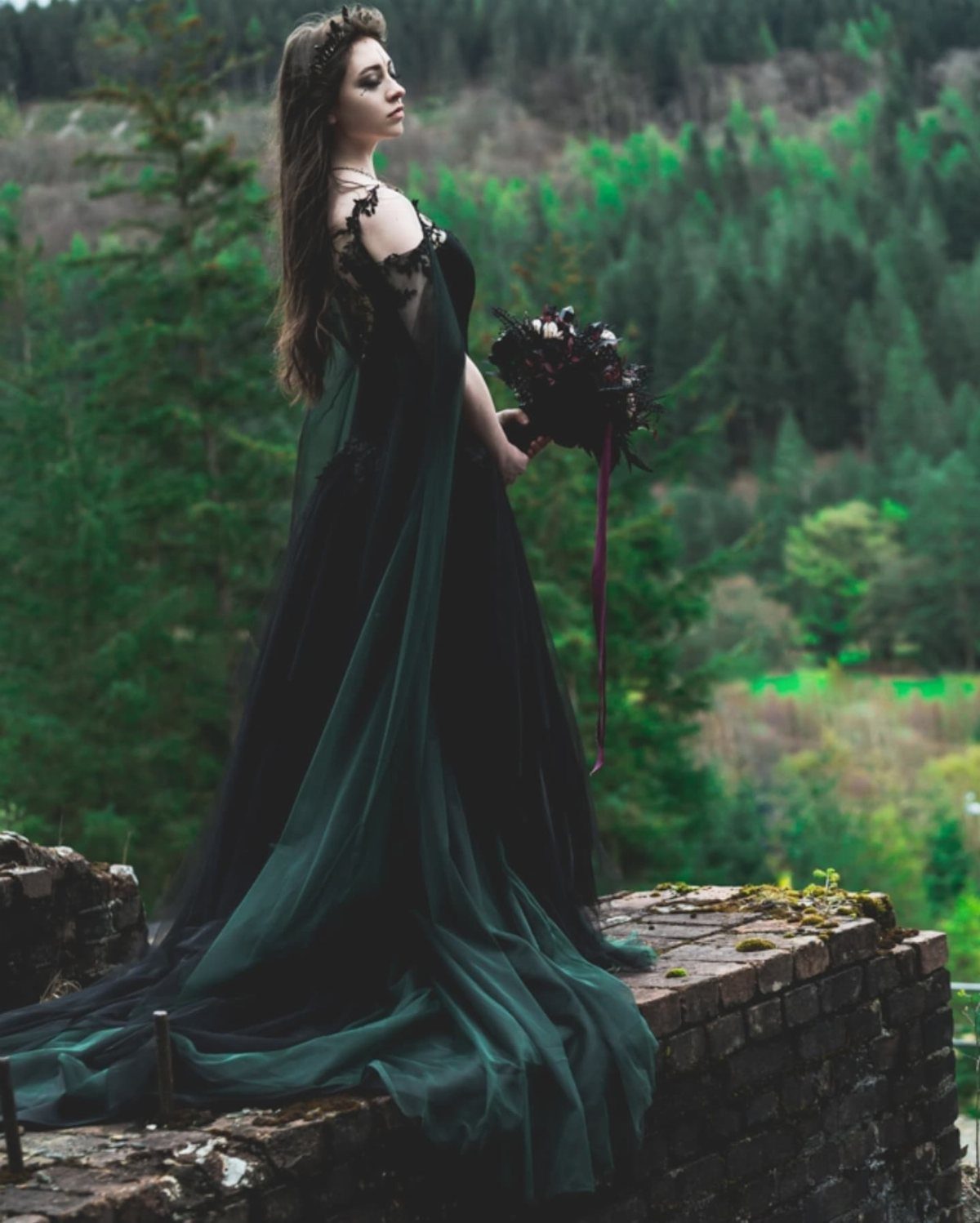A woman in a dark green tulle gown holding a bouquet in a forest.