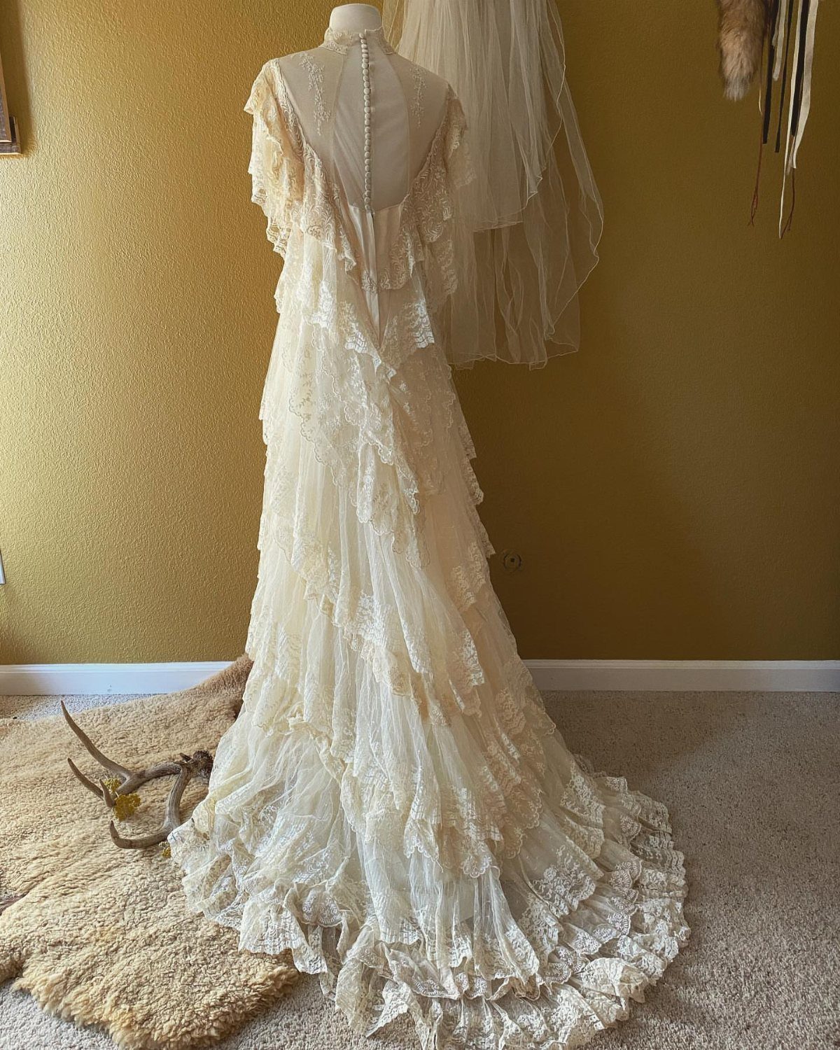 Ivory lace wedding dress with ruffled tiers displayed on a mannequin against a yellow wall.