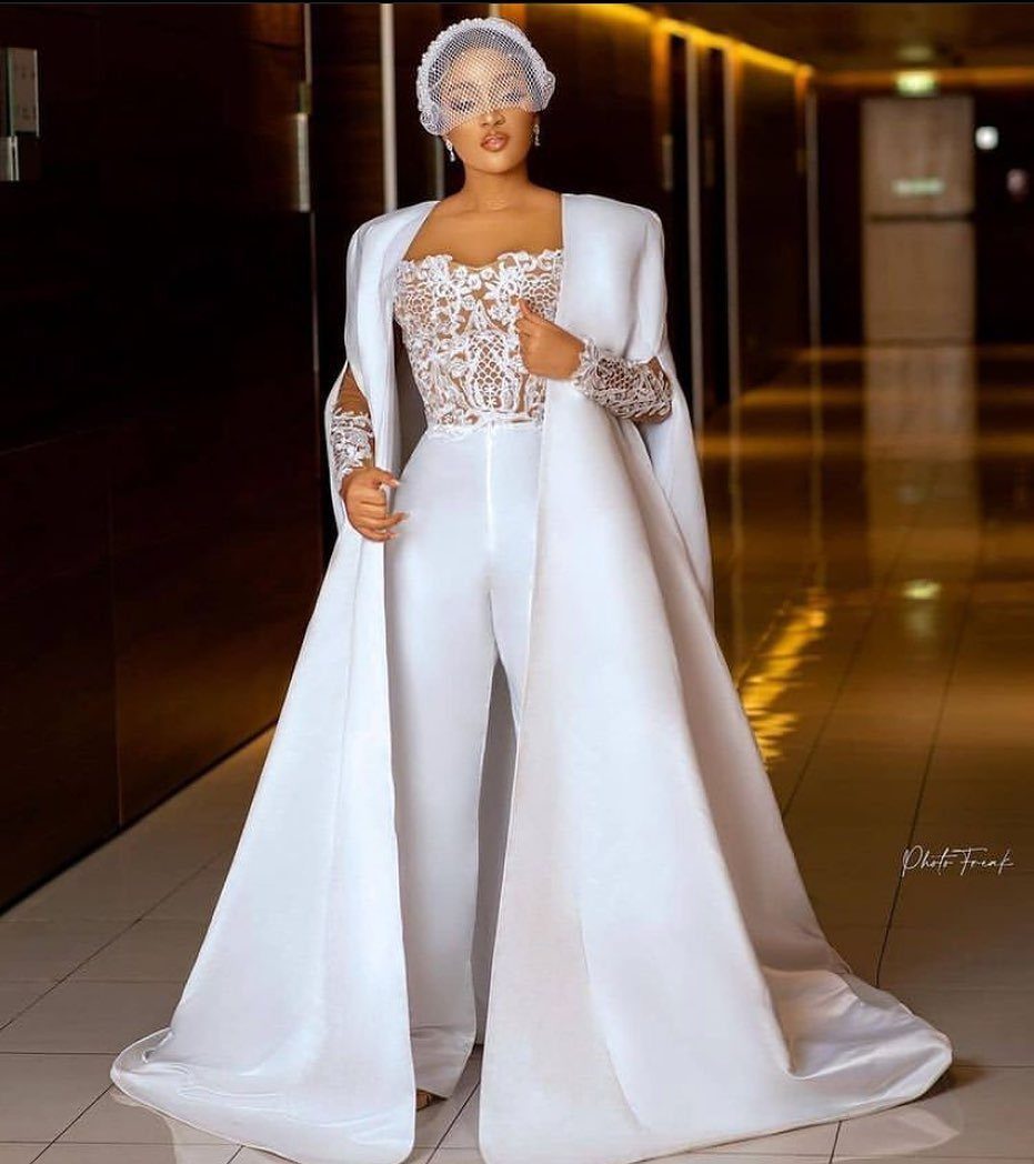 Woman wearing a white lace corset, wide-leg trousers, and a long flowing bridal cape.