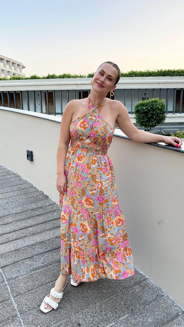 Woman in a colorful floral halter maxi dress and white sandals standing on a concrete ledge.