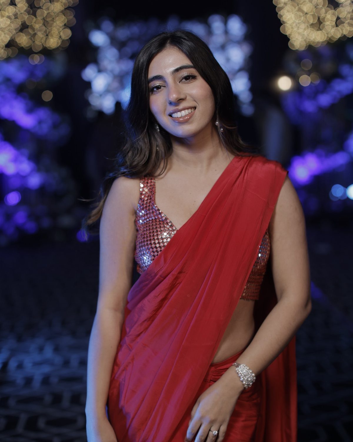 A smiling woman wearing a red saree and a sequined silver blouse against a blurred background.