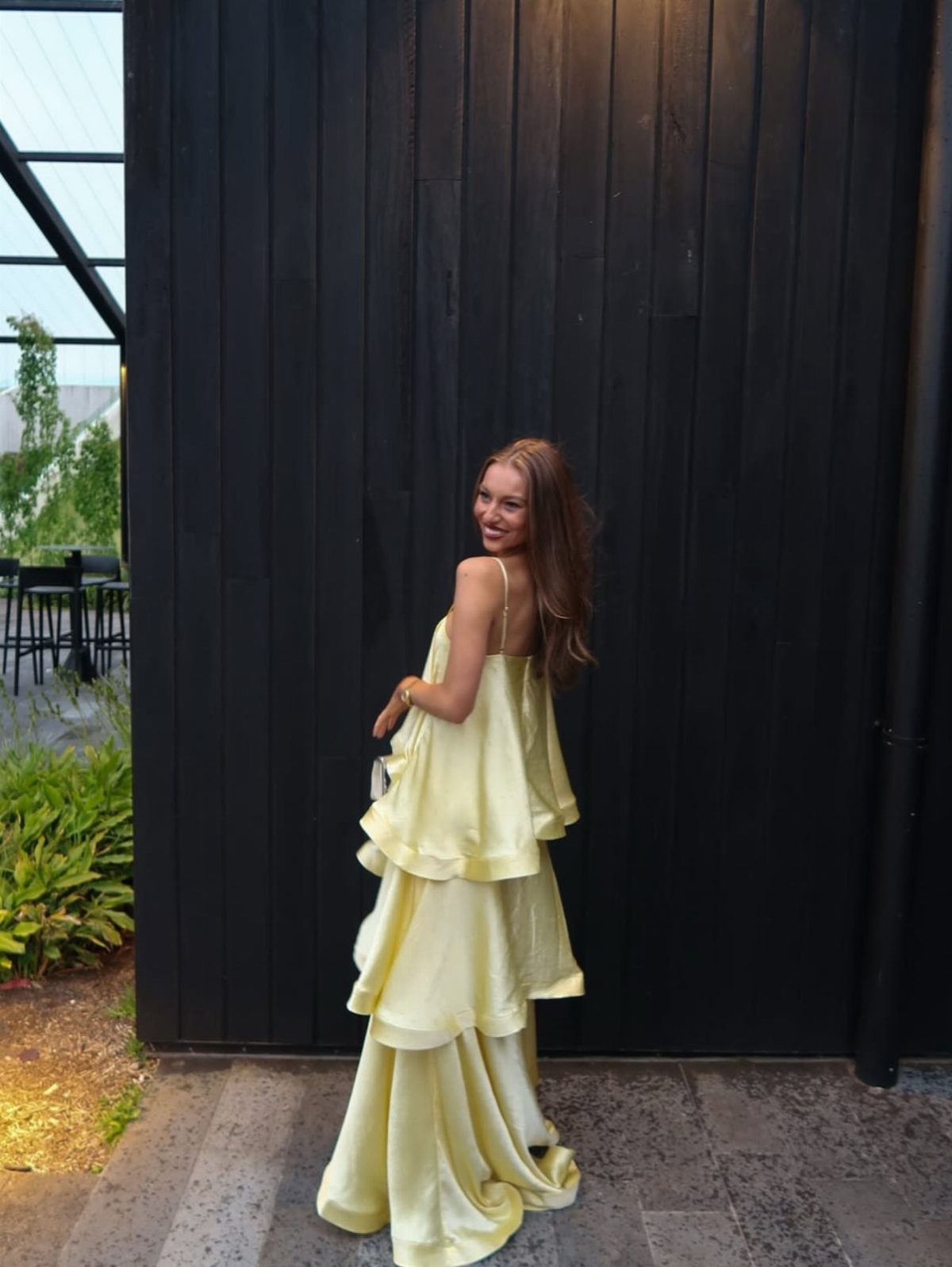 A woman in a tiered pale yellow satin dress posing against a dark wooden wall.