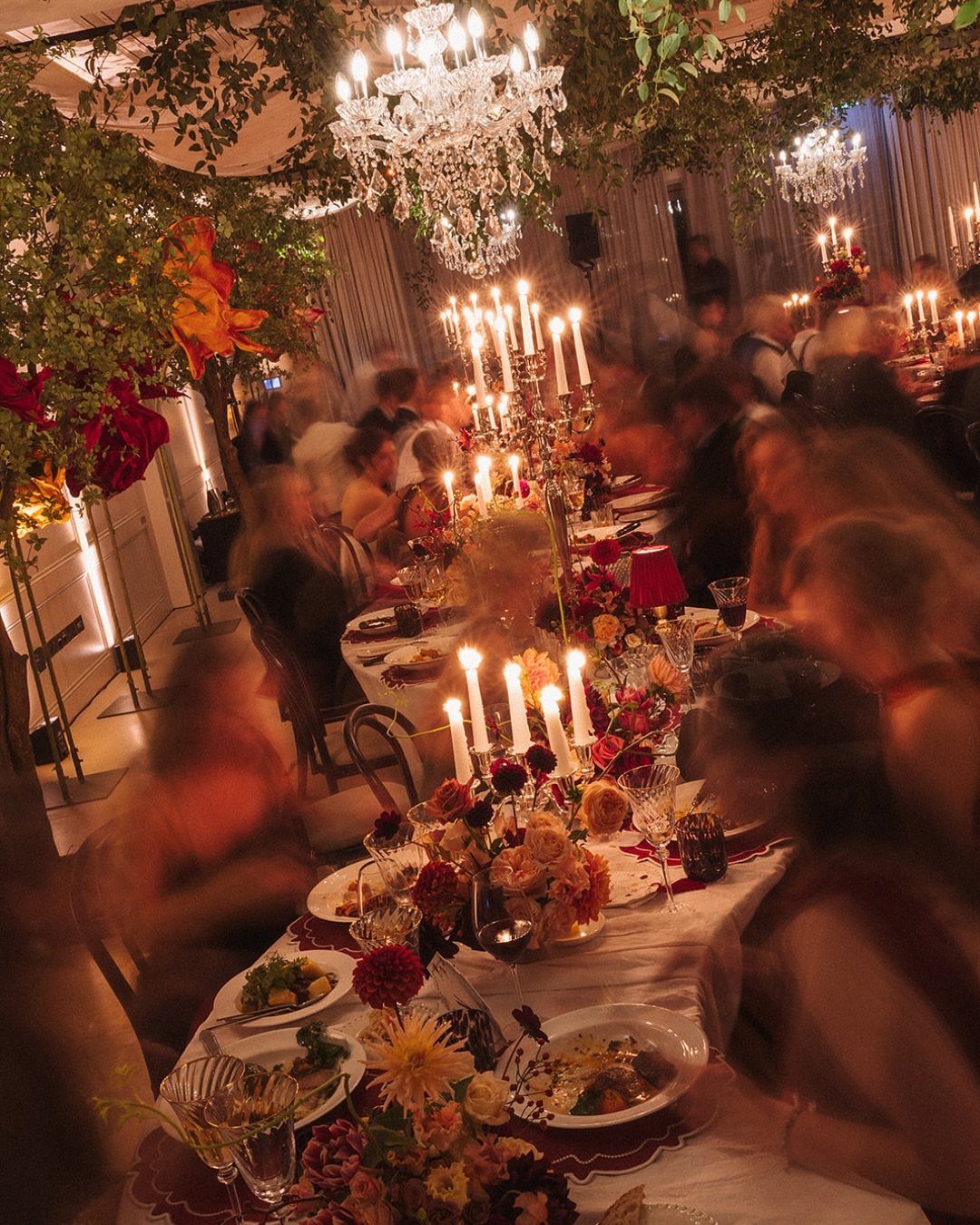 An elegantly set long dining table with warm candlelight, floral arrangements, and crystal chandeliers.