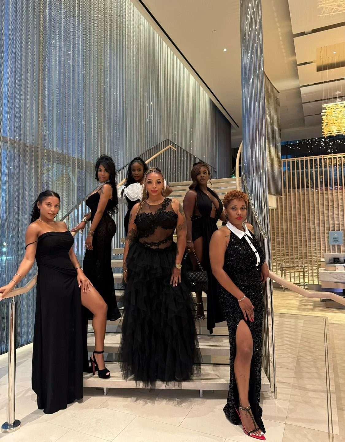 Six women in elegant black formal gowns posing on a staircase in a modern building.