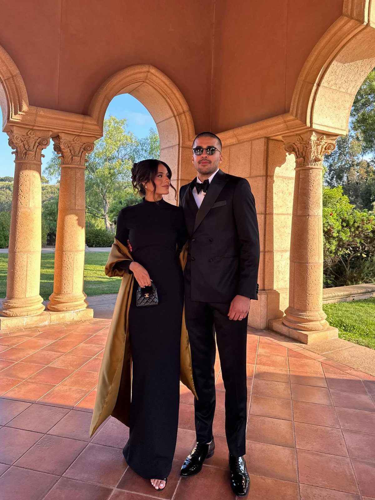 A man in a tuxedo and a woman in a black gown posing under stone arches.