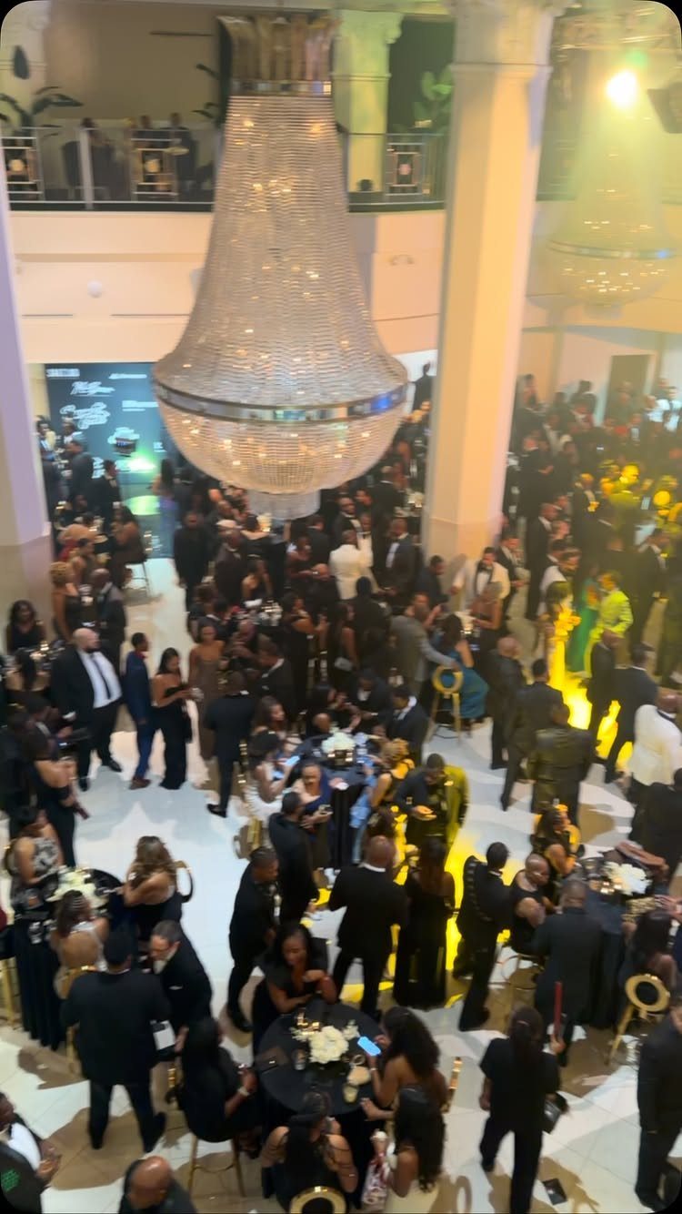 Crowded ballroom with people in formal black attire under a large crystal chandelier.