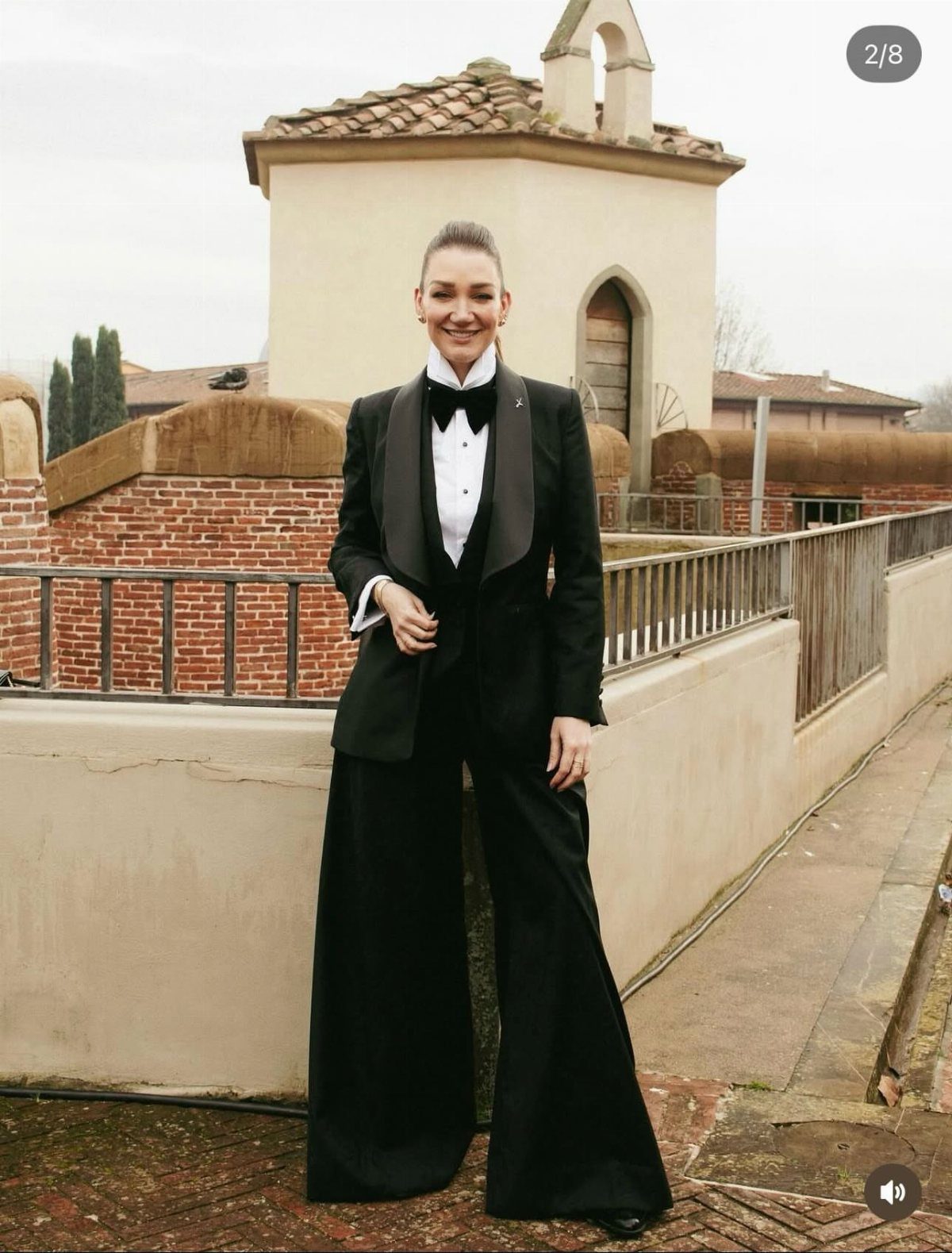 A woman posing in a black satin lapel tuxedo and wide-leg trousers on a terrace.