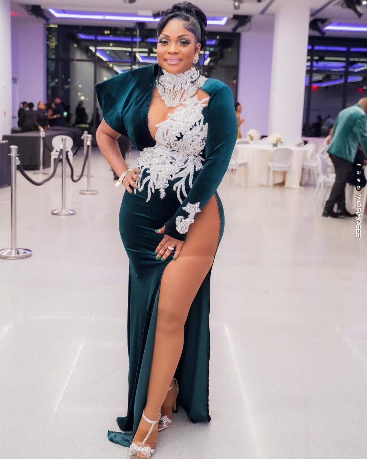 Woman in an emerald green velvet gown with white floral embroidery and a high leg slit.