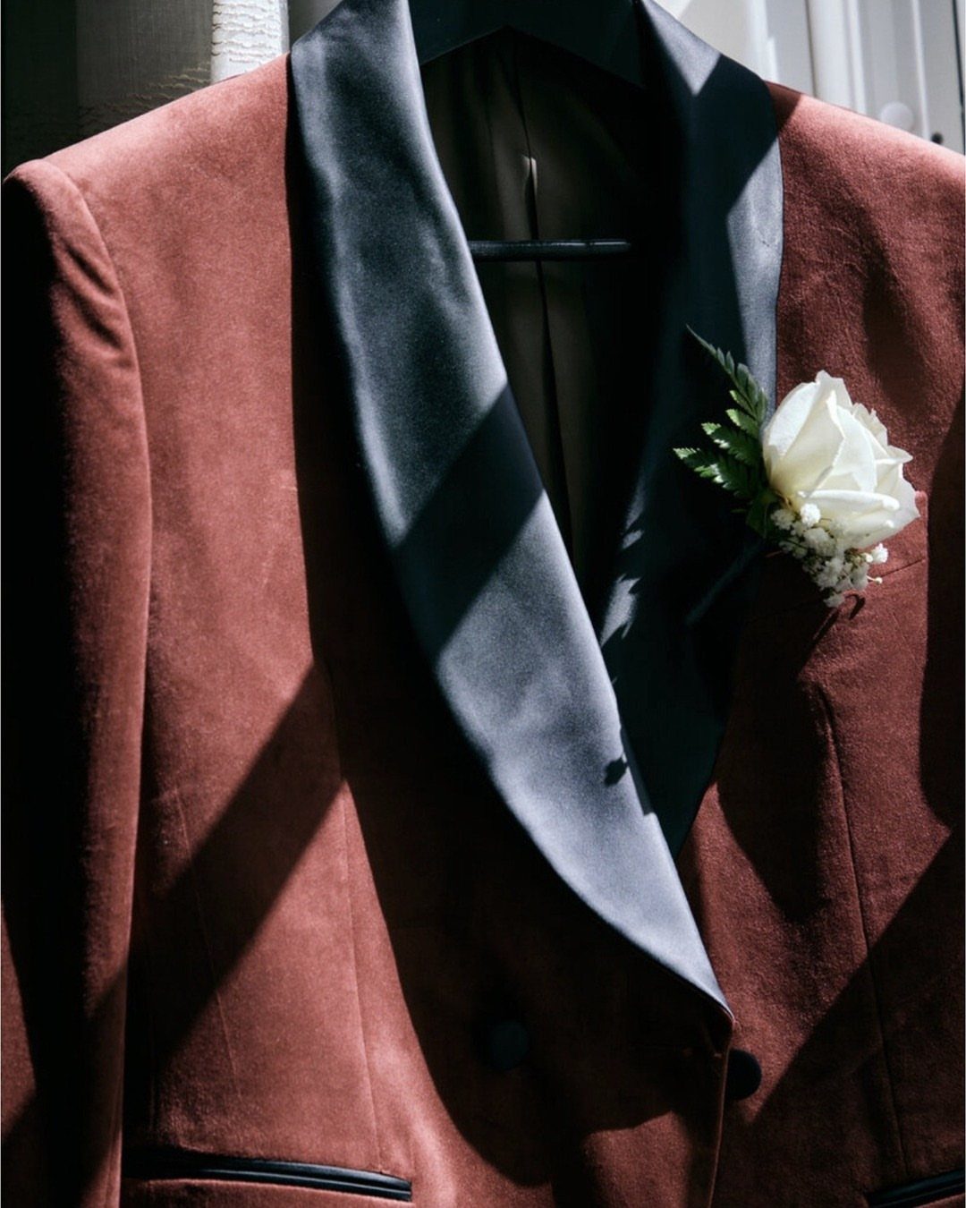 A rust-colored velvet tuxedo jacket featuring black satin lapels and a white rose boutonniere.