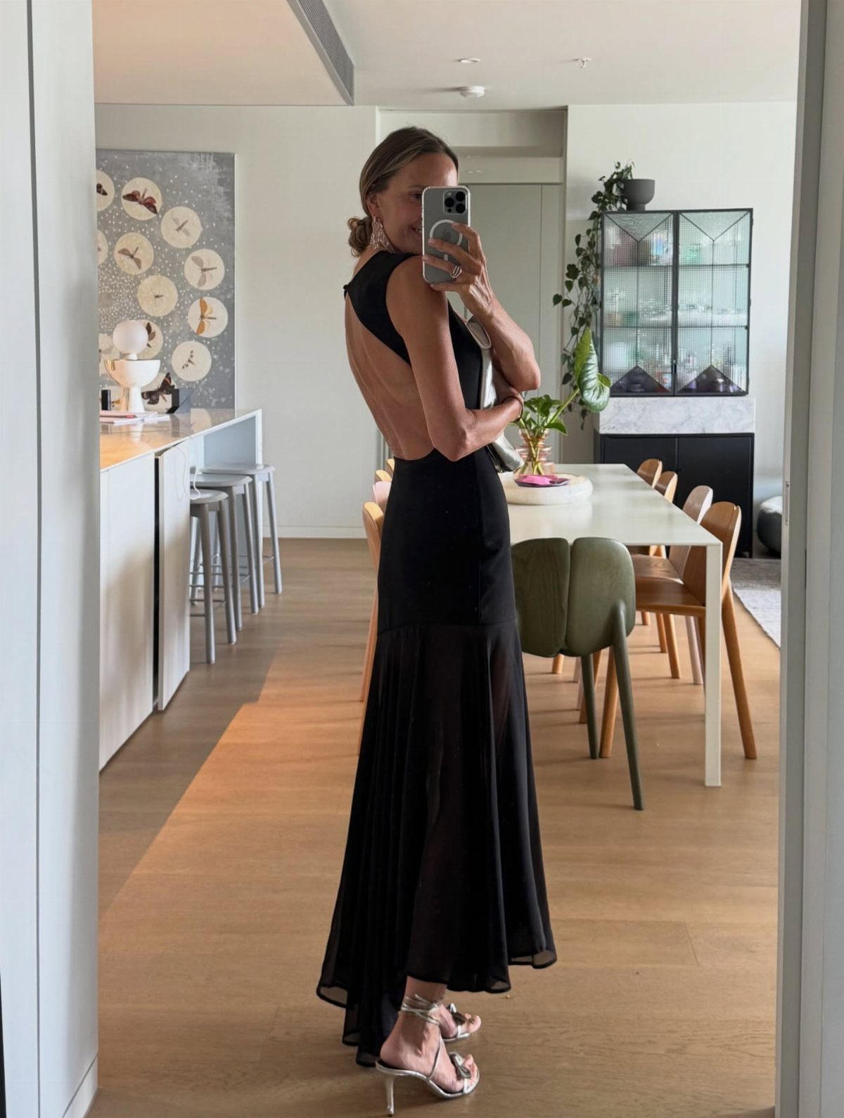 Woman in a black backless maxi dress and silver heels posing for a mirror selfie.