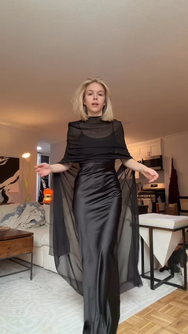 Blonde woman posing in a formal floor-length black satin gown with a sheer cape overlay.