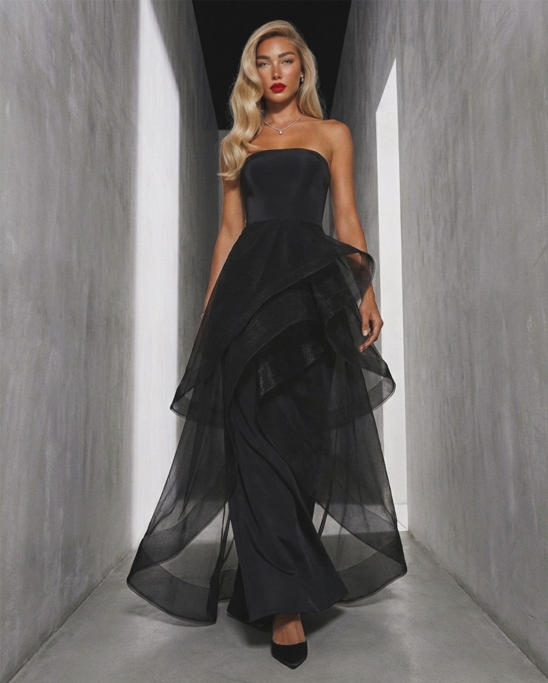 A blonde woman posing in a formal strapless black gown within a concrete hallway.