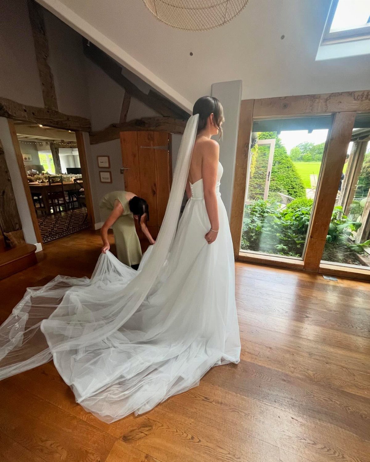 Bride in a white gown standing while a bridesmaid adjusts her long veil and train.