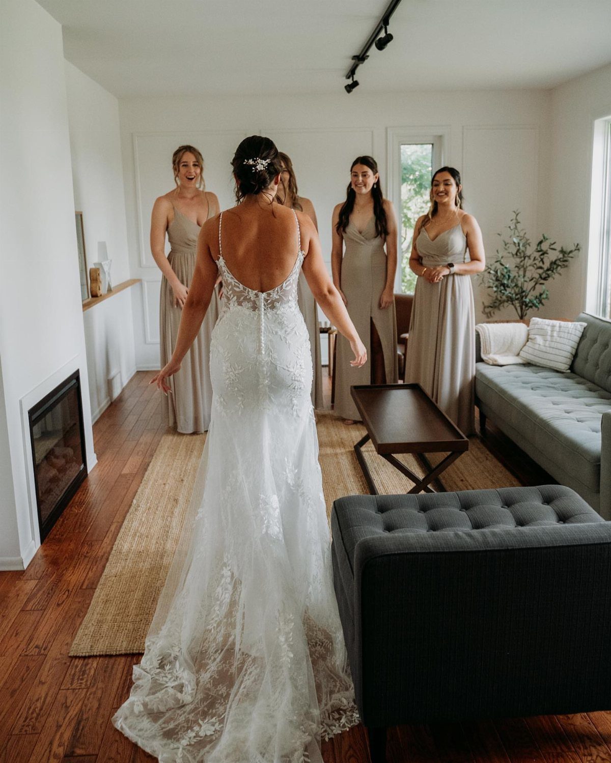 Bride in a white lace gown facing her bridesmaids who are reacting with smiles in a living room.