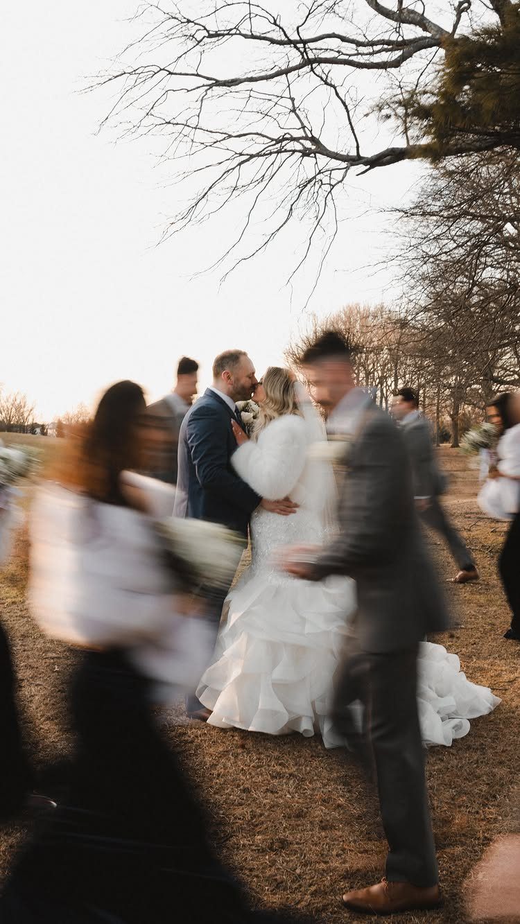 A bride and groom kissing while blurred wedding guests move around them in a field.