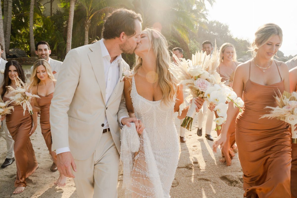Bride and groom kissing while walking on a beach surrounded by their wedding party.