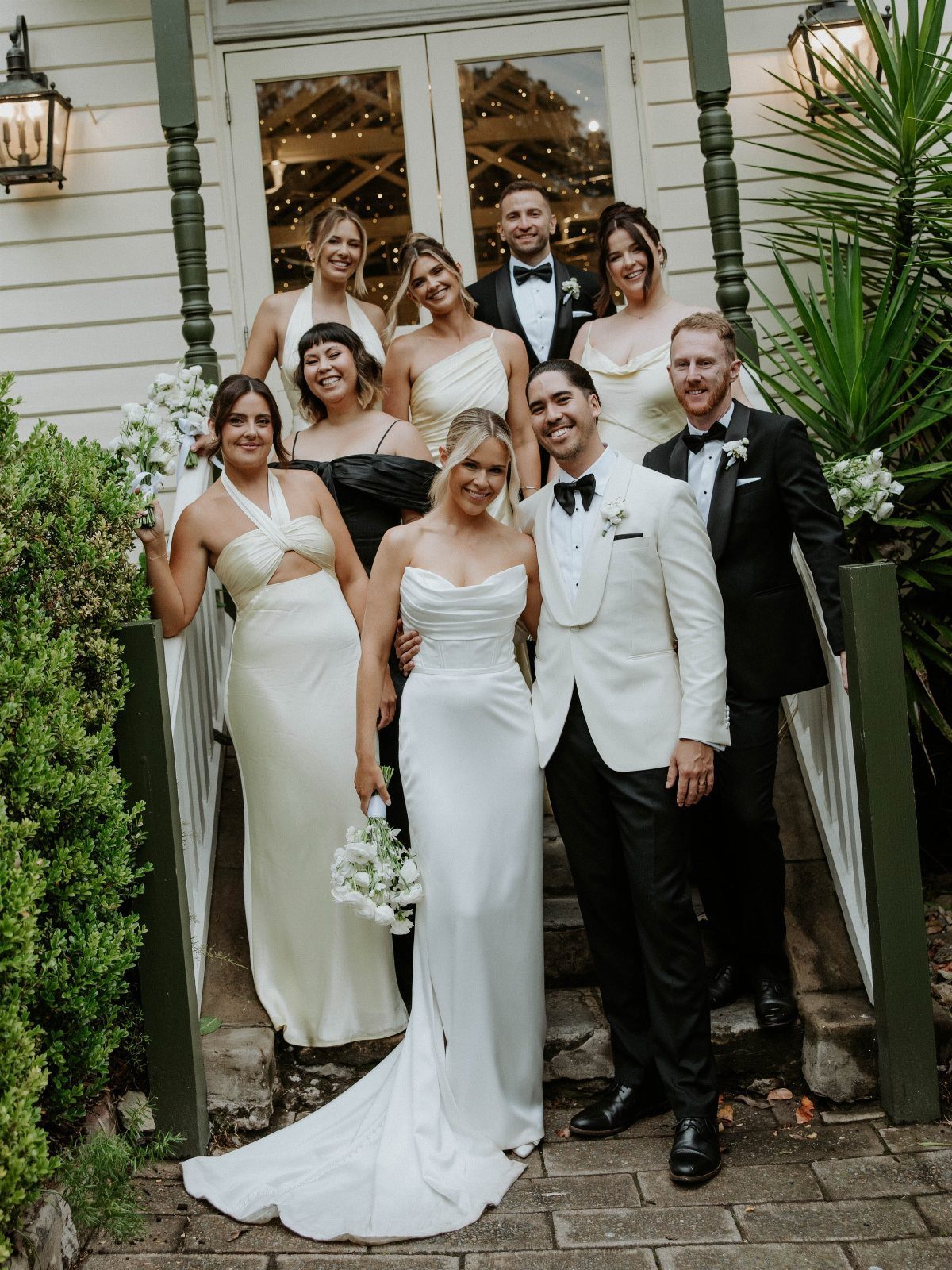 A wedding party posing together on the steps of a white house with green trim.