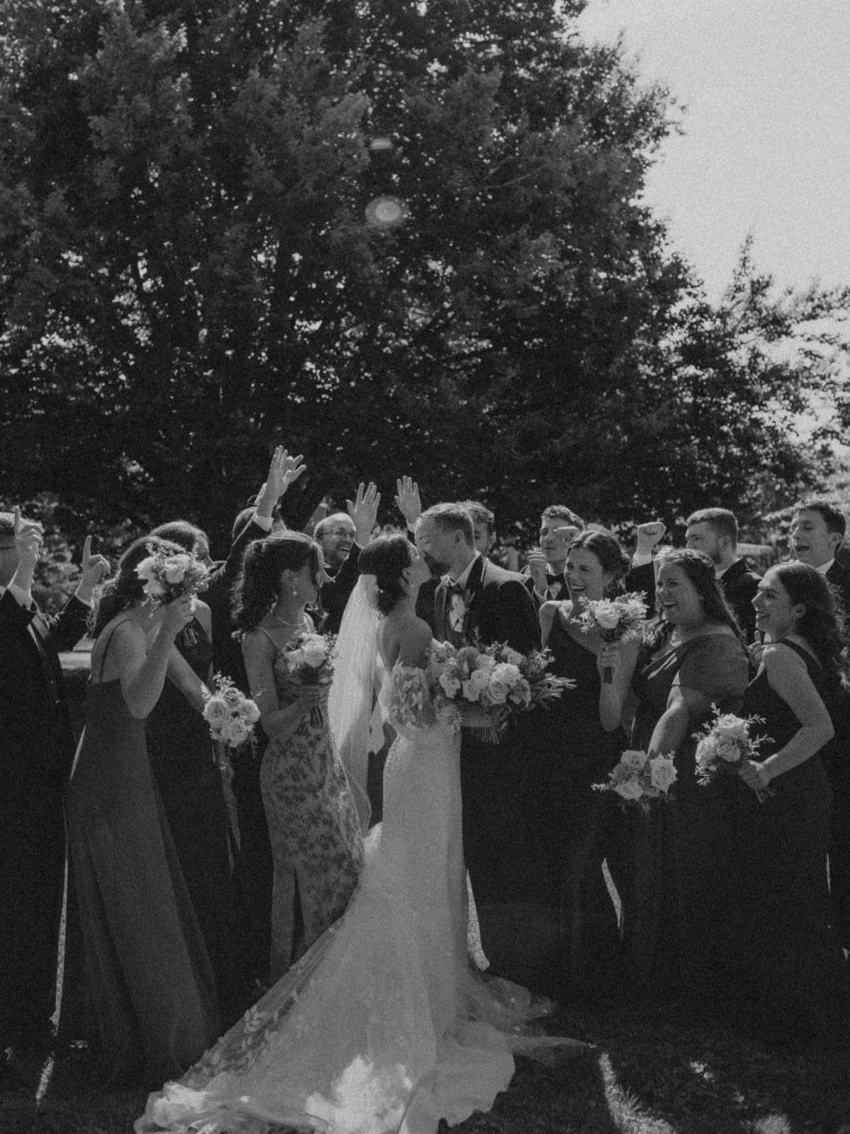 A bride and groom surrounded by their cheering bridal party in a celebratory outdoor setting.