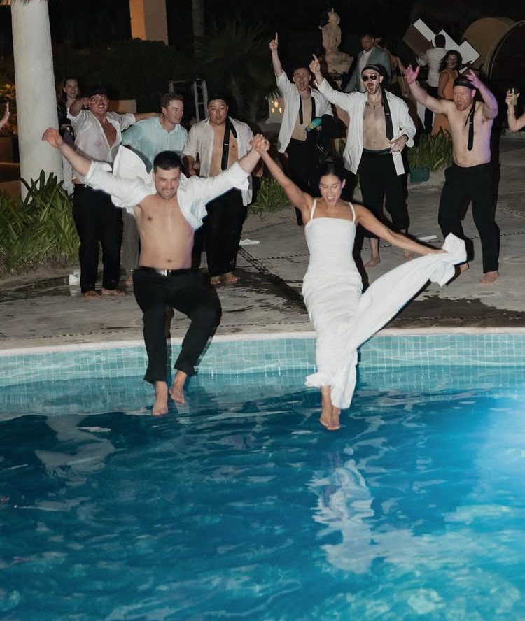 A bride and groom jumping into a blue swimming pool at night surrounded by cheering wedding guests.