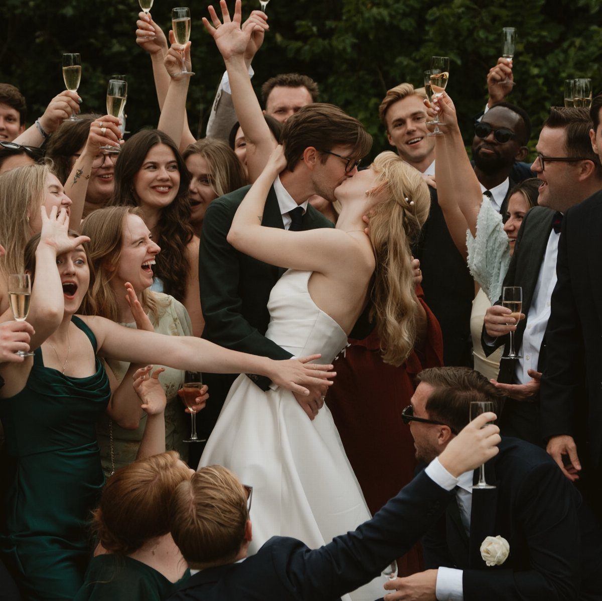 A bride and groom kiss while surrounded by a cheering wedding party holding champagne glasses.