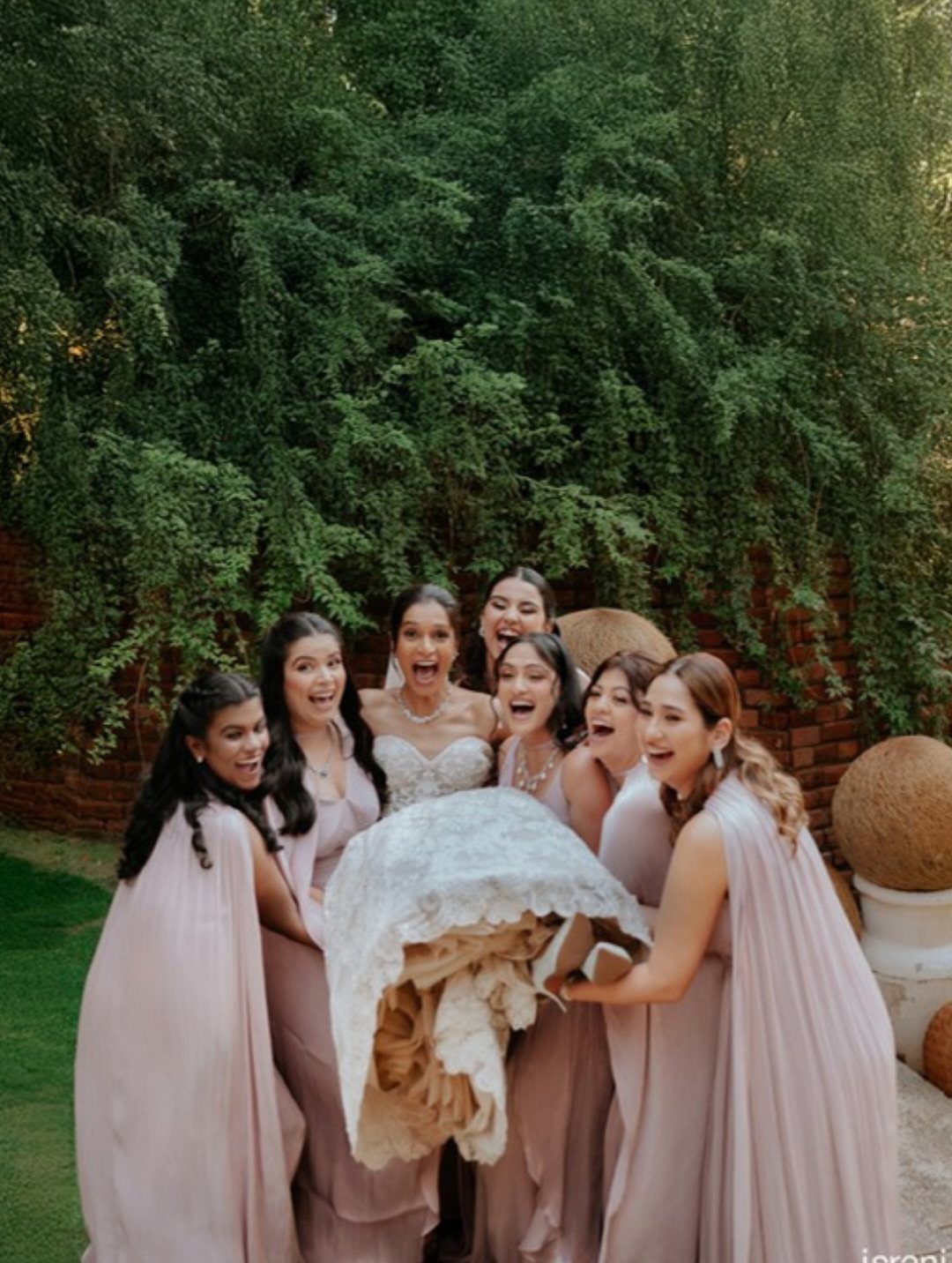 Bridesmaids in pink dresses lifting a bride in a white lace gown outdoors.