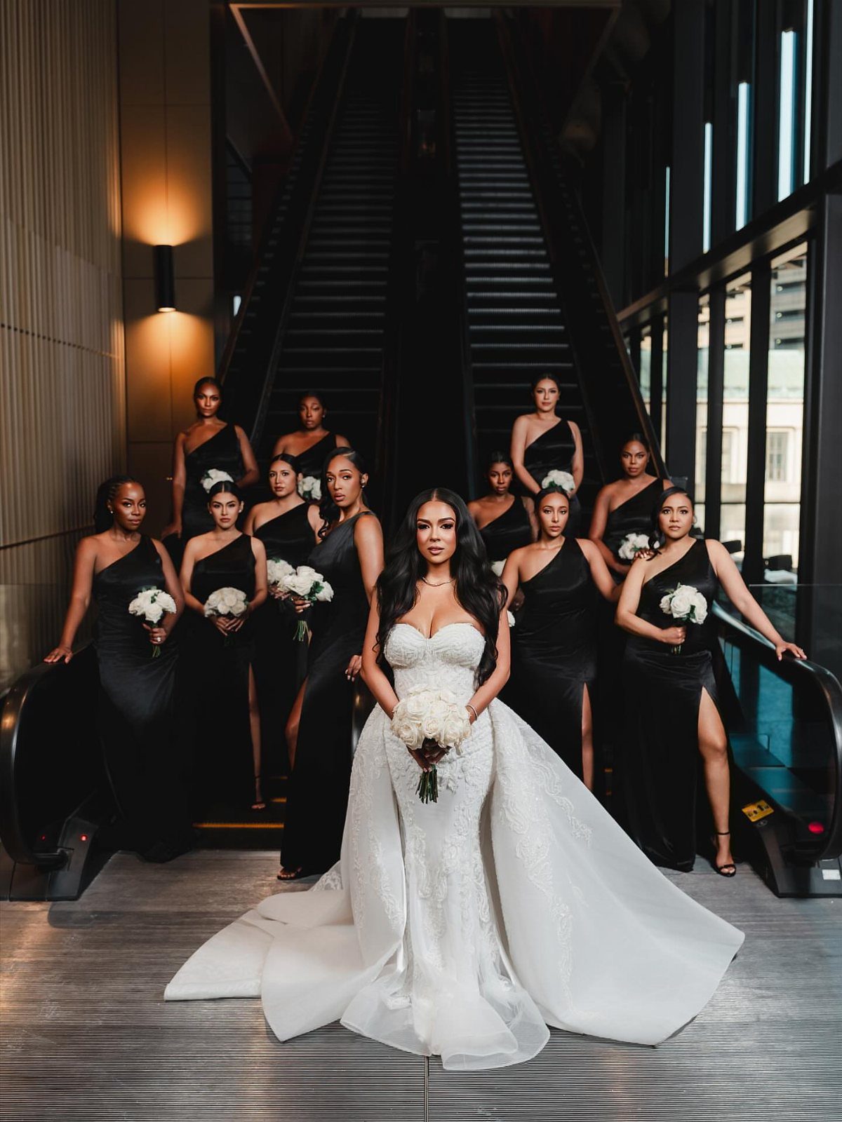 Bride in white gown surrounded by bridesmaids in black dresses holding white bouquets on escalators.