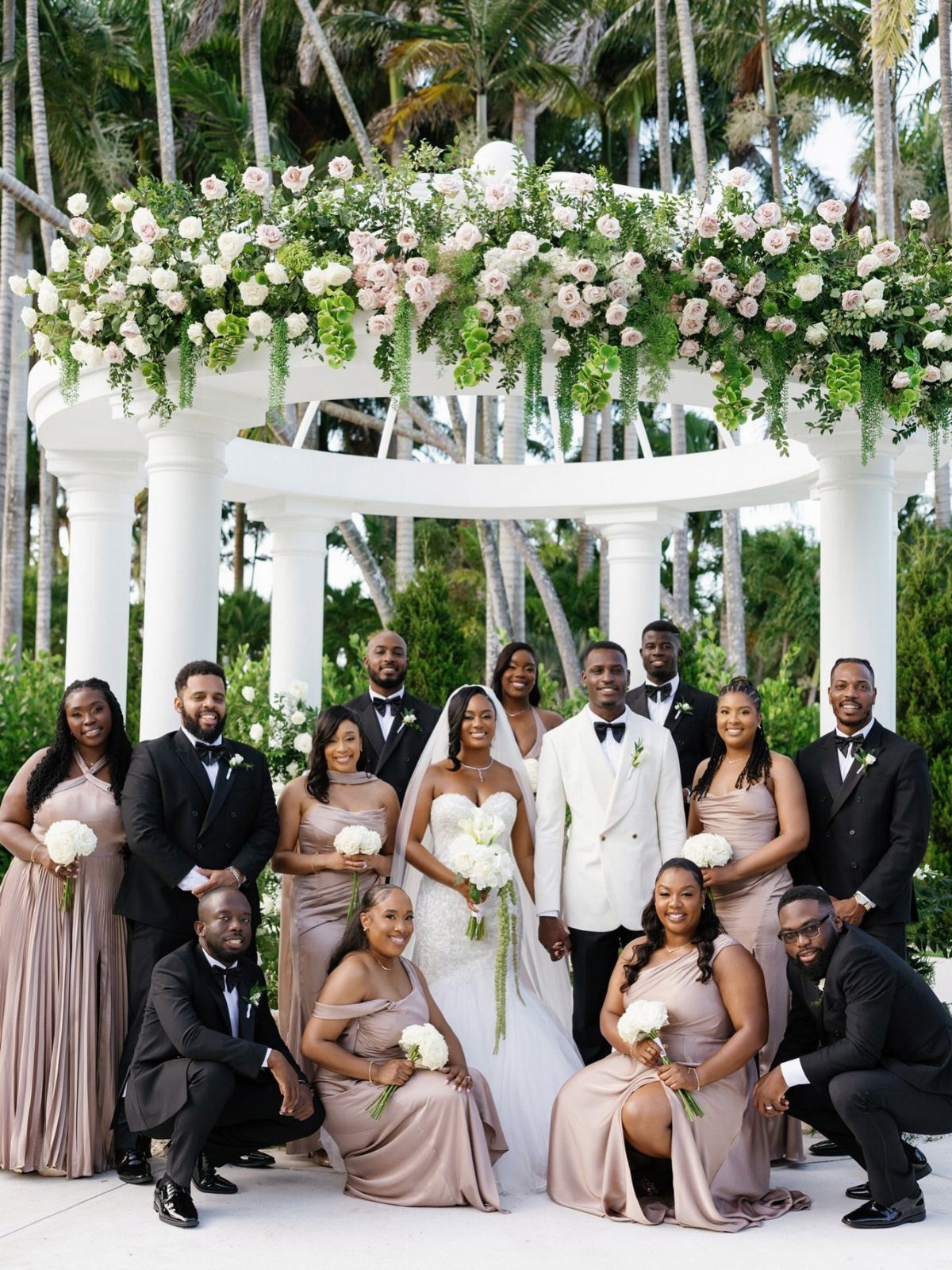 A large wedding party posing together in front of a white floral gazebo and palm trees.