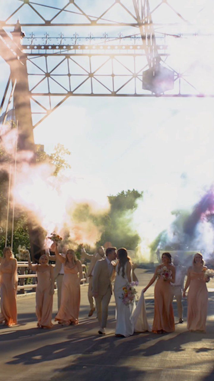 Bridal party walking through colorful smoke bombs under a large metal bridge structure.