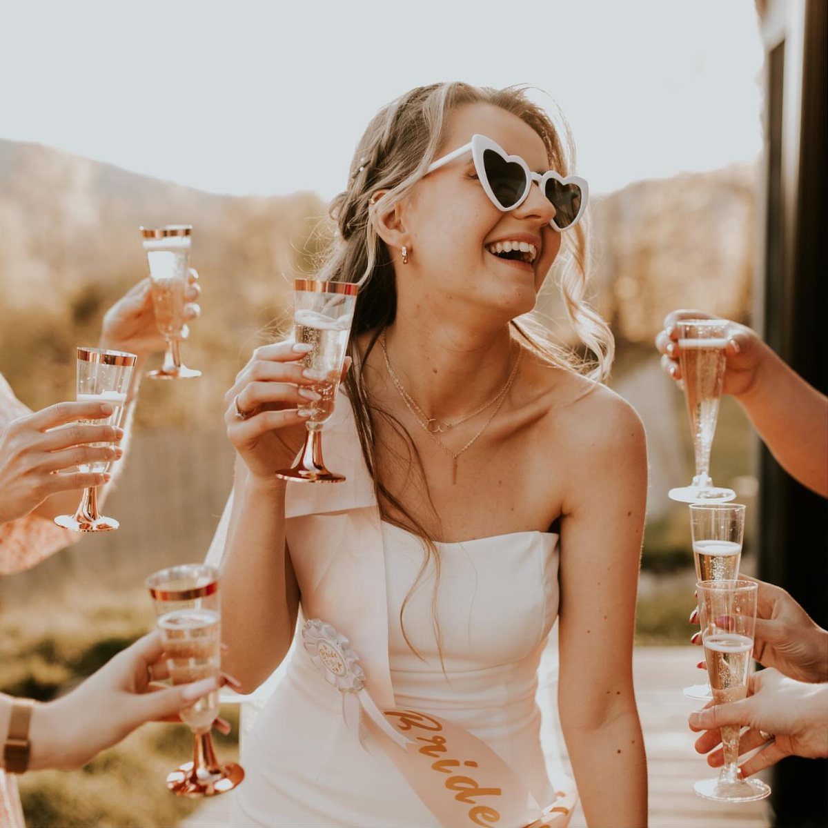 A laughing bride in heart sunglasses surrounded by hands holding champagne glasses.