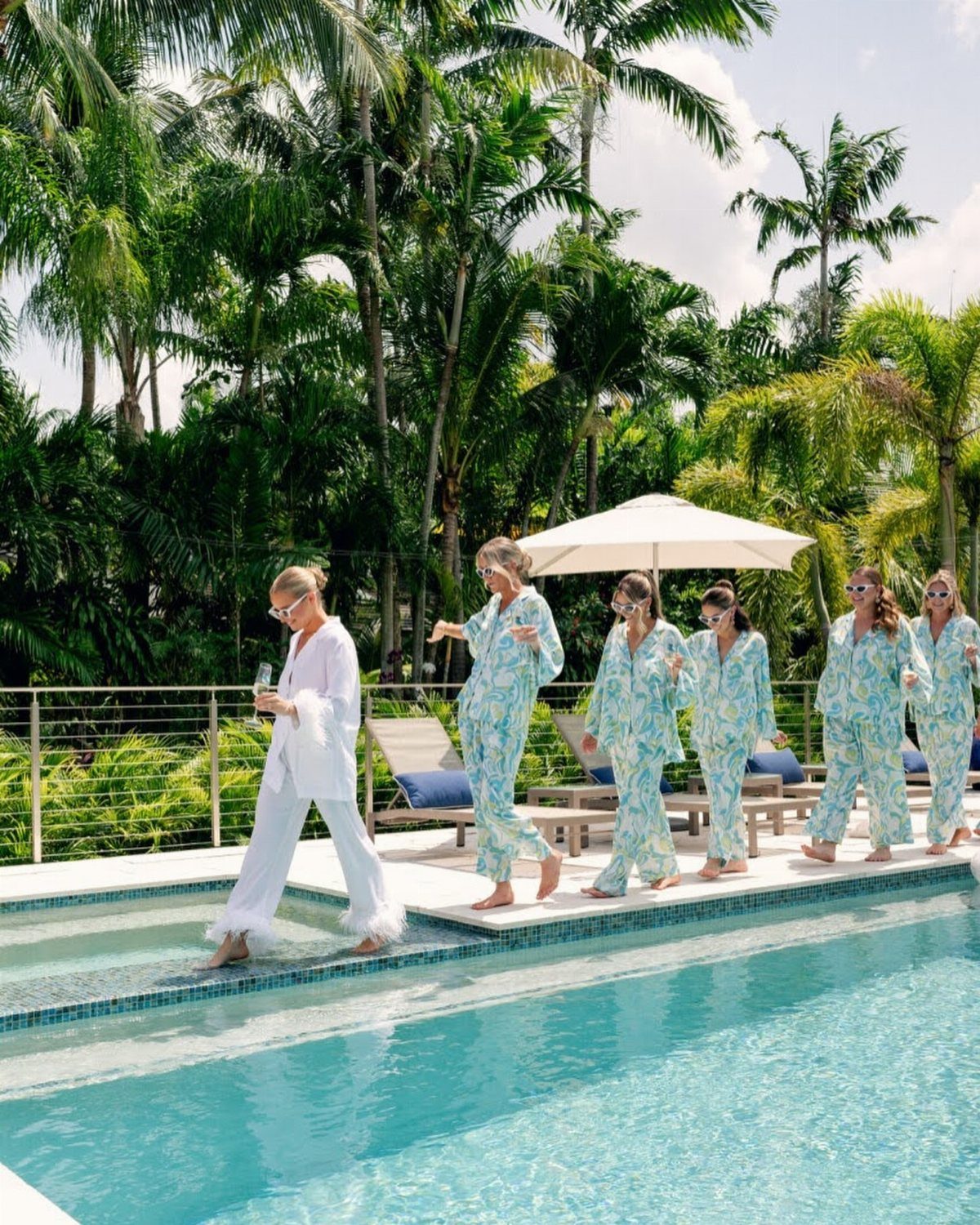 Women in matching patterned pajamas walking in a line beside a tropical swimming pool.