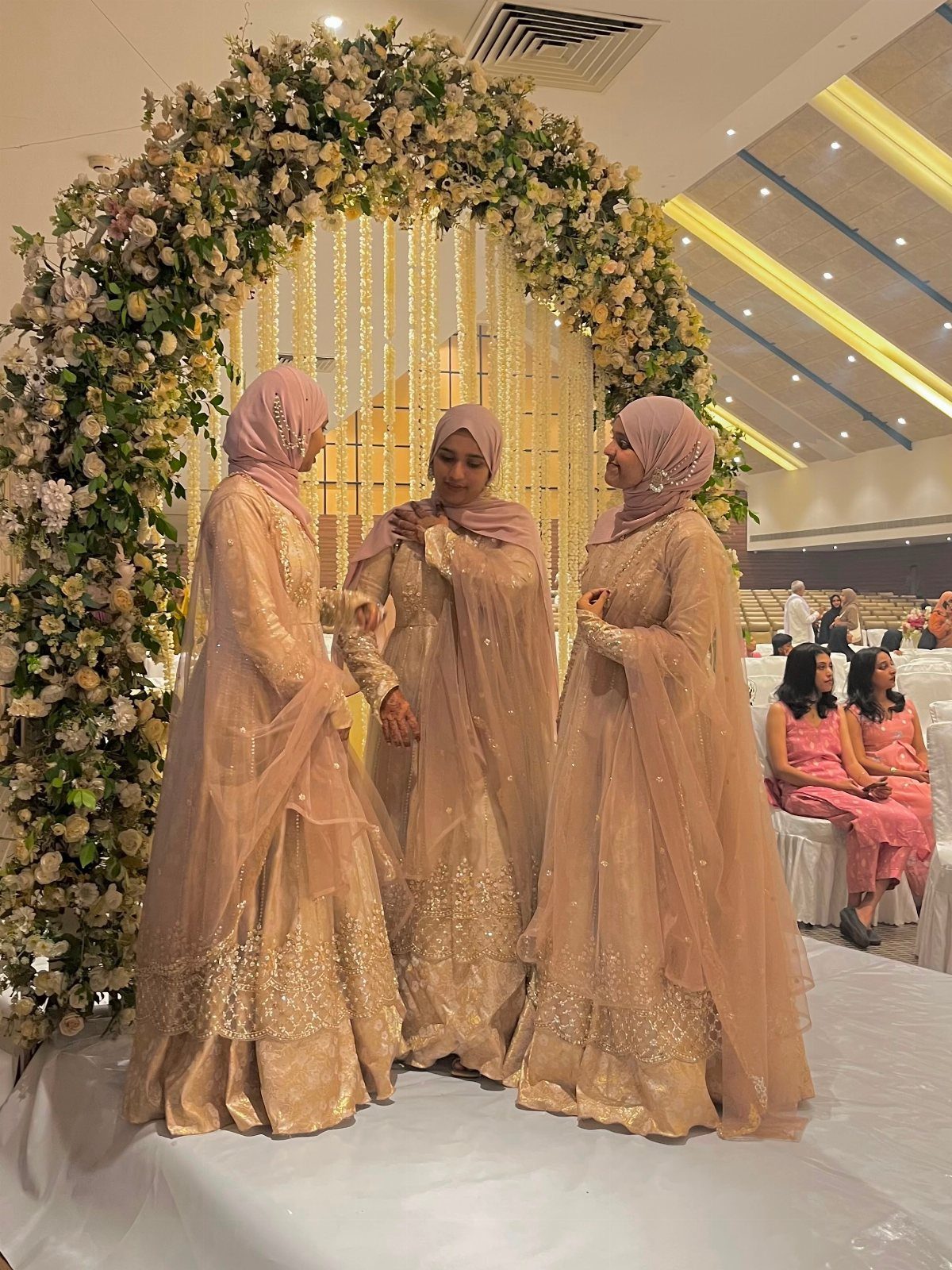 Three women in matching peach embellished dresses and hijabs standing under a floral arch.