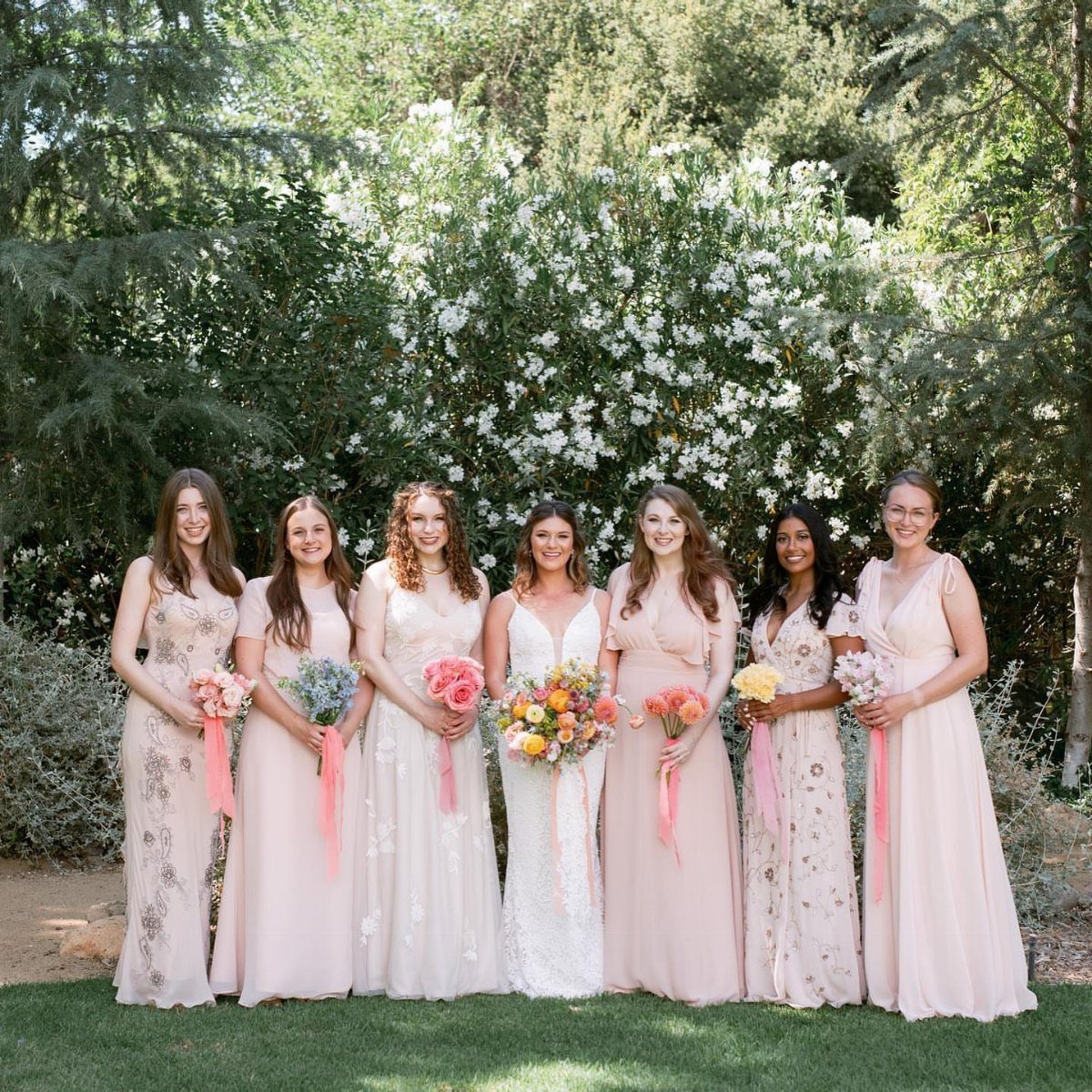 Seven bridesmaids in various shades of pink and cream dresses holding colorful bouquets outdoors.