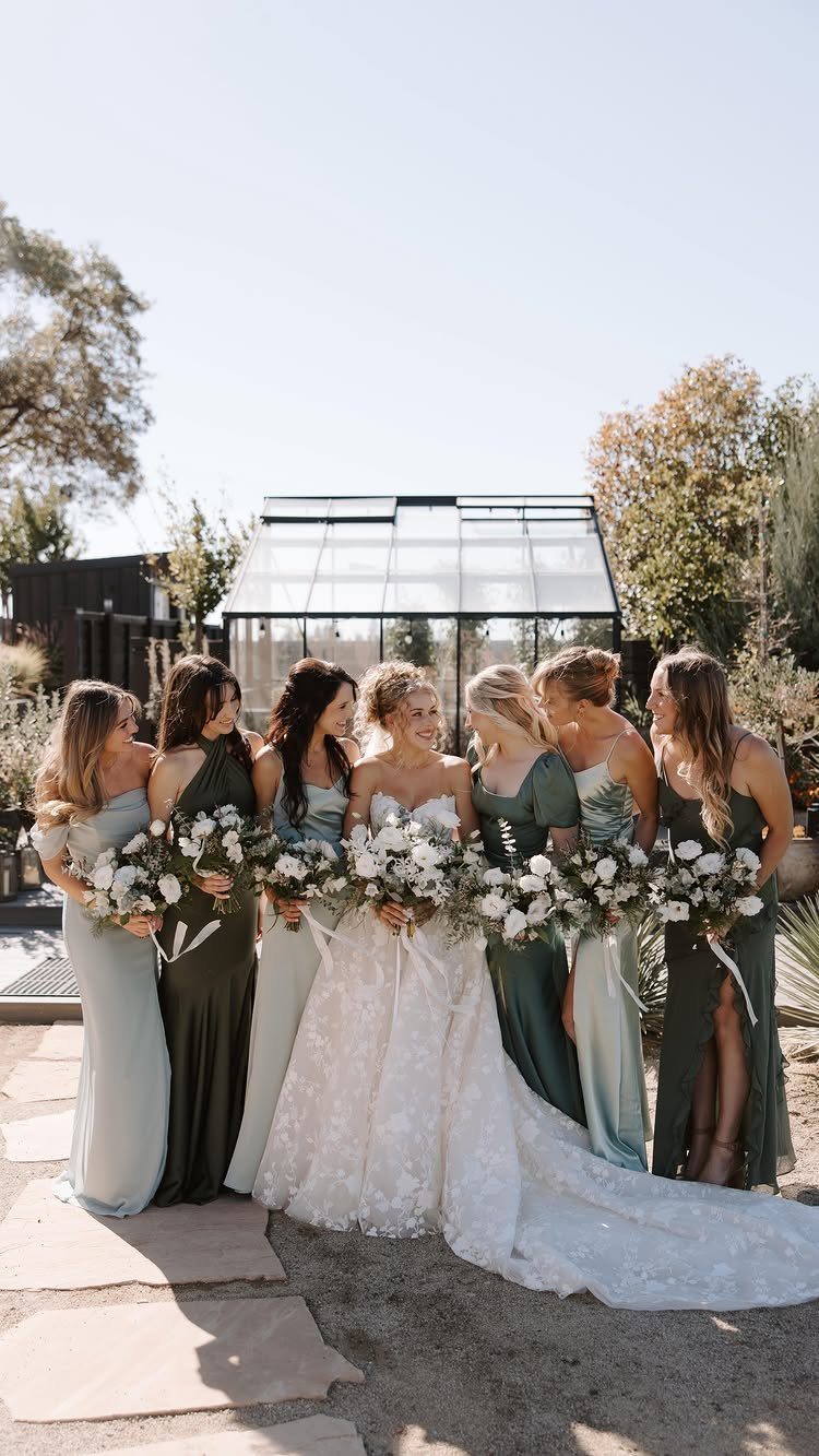 Bridesmaids in long green and blue dresses standing with a bride in a white lace gown.