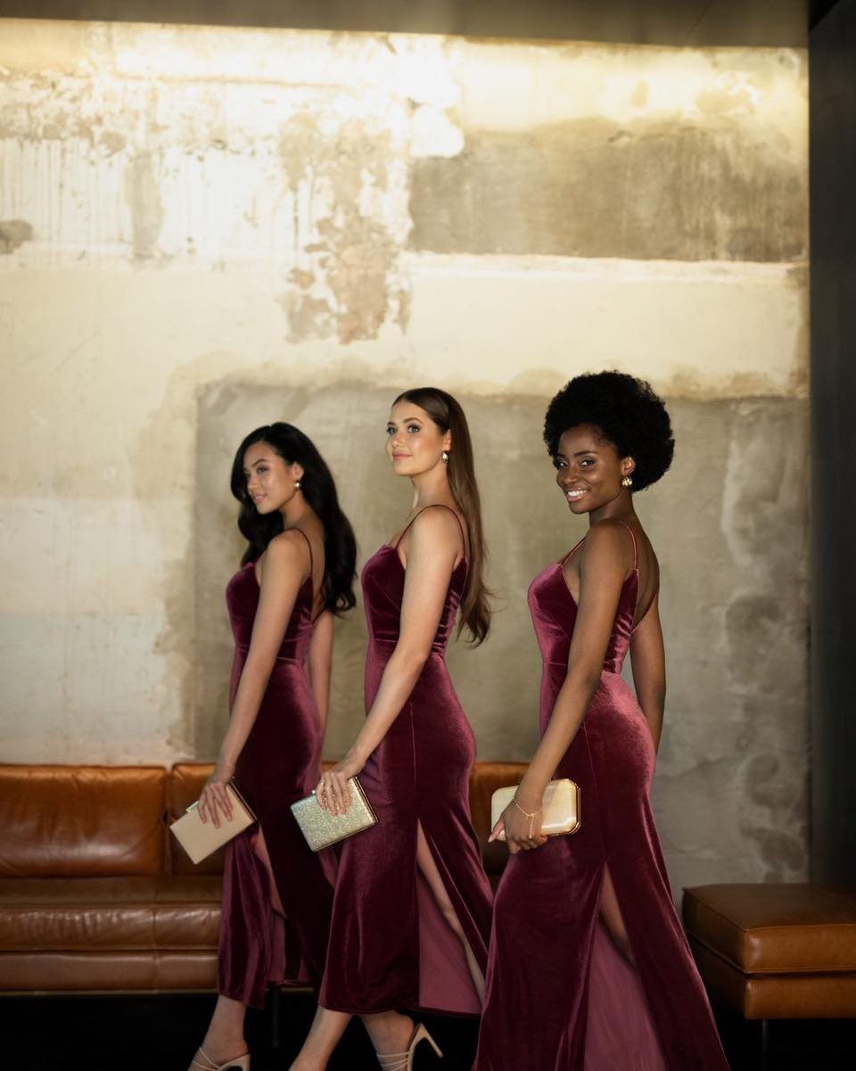 Three women posing in long burgundy velvet dresses while holding gold clutches.