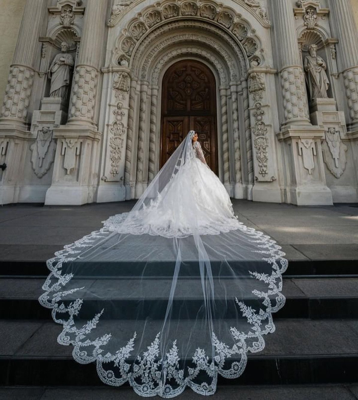 Bride in a white gown wearing a long lace-edged cathedral veil on cathedral steps.