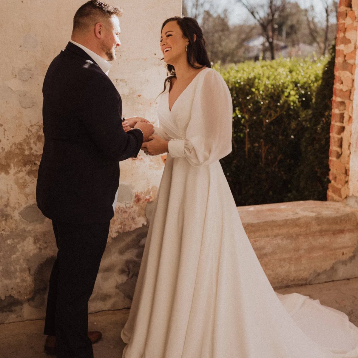 Bride in a white long-sleeved gown with bishop sleeves smiling at her groom outdoors.