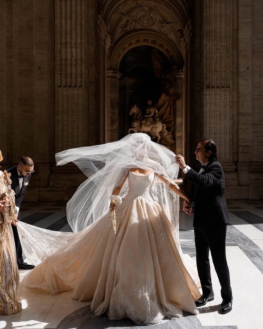 A bride in a voluminous embroidered ballgown and long veil being assisted by two men.