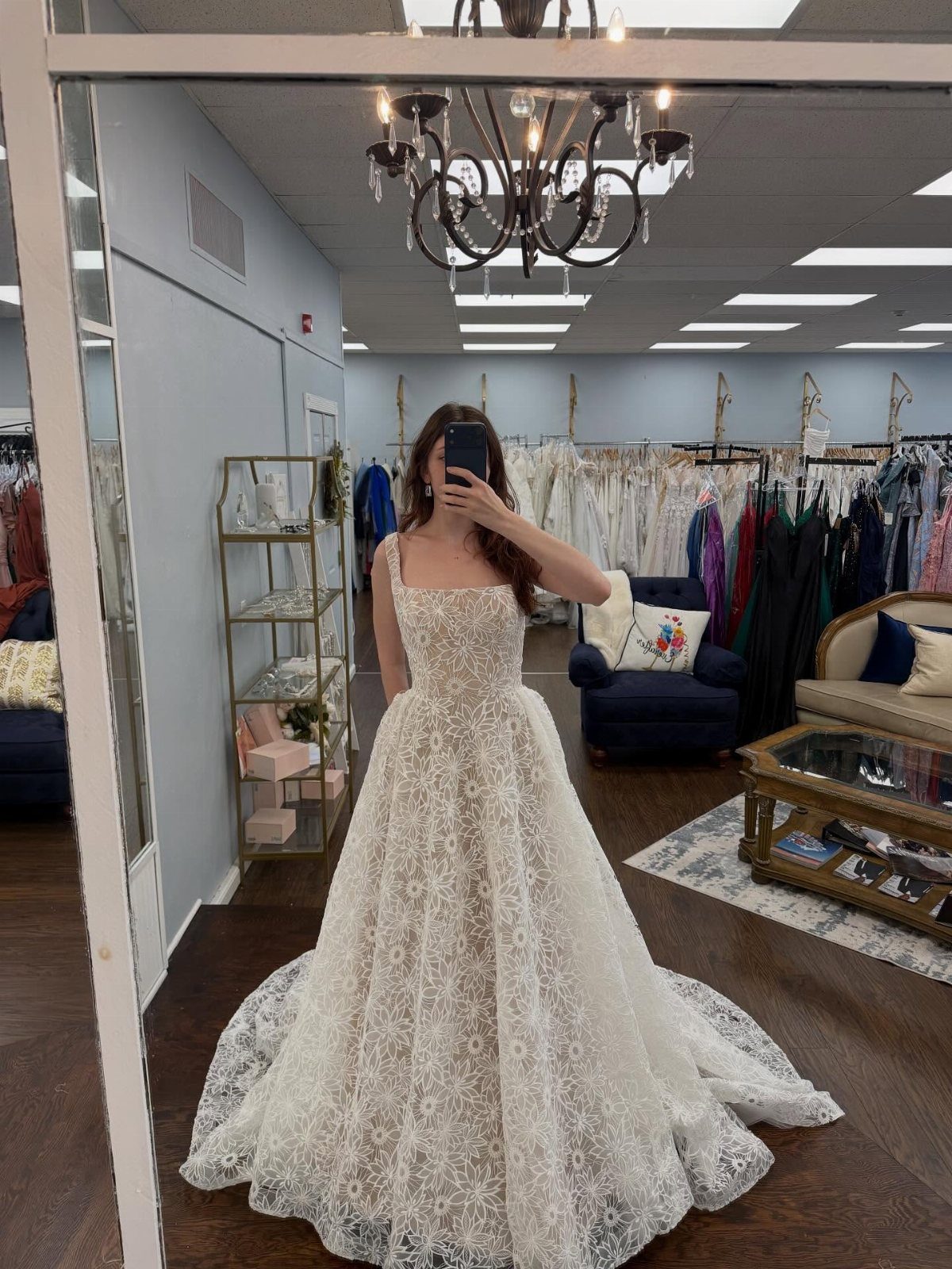 Woman in a white lace A-line wedding dress taking a mirror selfie in a boutique.