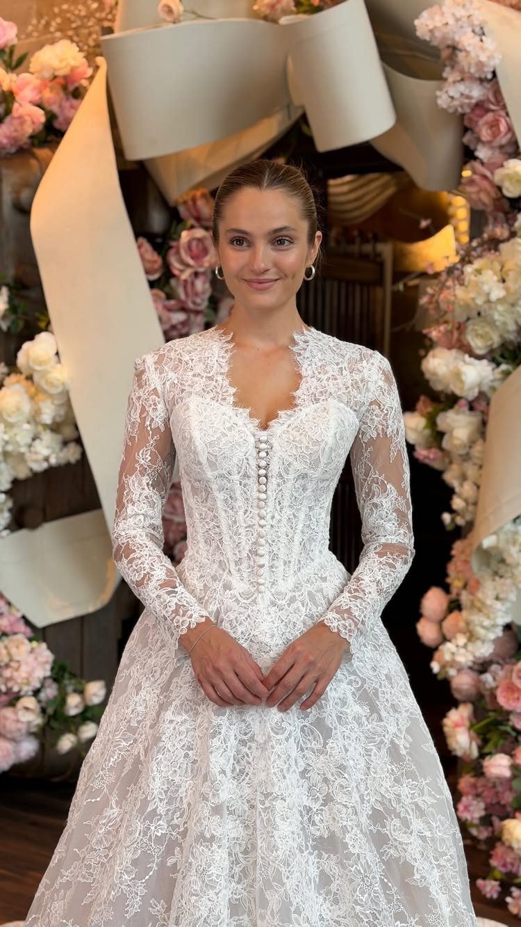 Woman in a white long-sleeved lace wedding dress posing amidst flowers and a large bow.