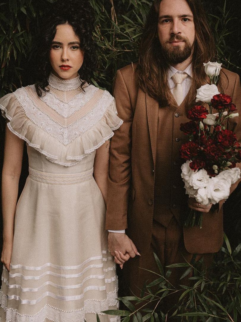 Woman in a vintage cream lace wedding dress standing with a man in a brown suit.