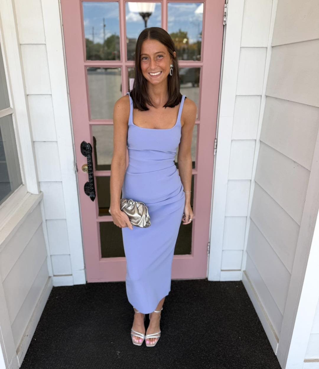A smiling woman in a lavender midi cocktail dress posing in front of a pink door.