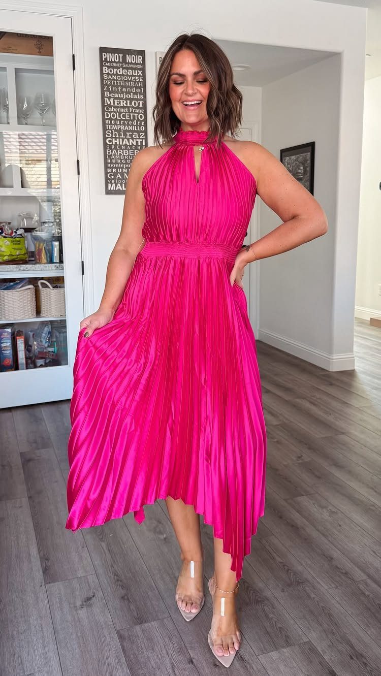 A woman smiling while wearing a bright pink pleated halter cocktail dress indoors.