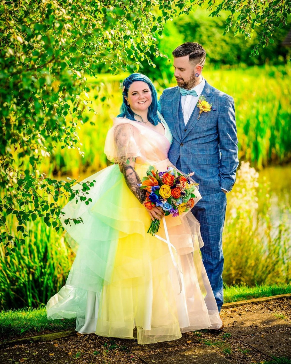 A bride in a pastel rainbow tulle wedding dress and a groom standing outdoors.