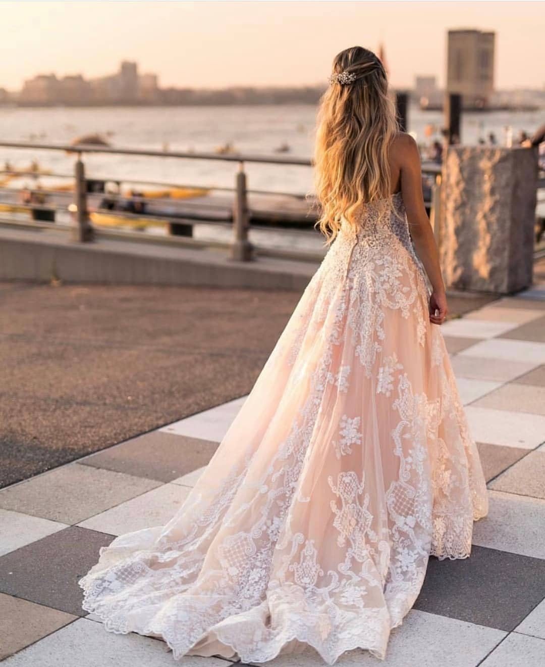 Woman in a blush lace wedding dress standing on a waterfront walkway during sunset.