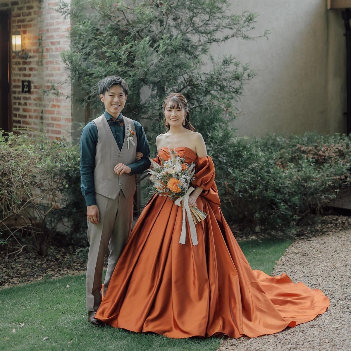 A bride in a burnt orange satin gown and a groom posing in a garden.