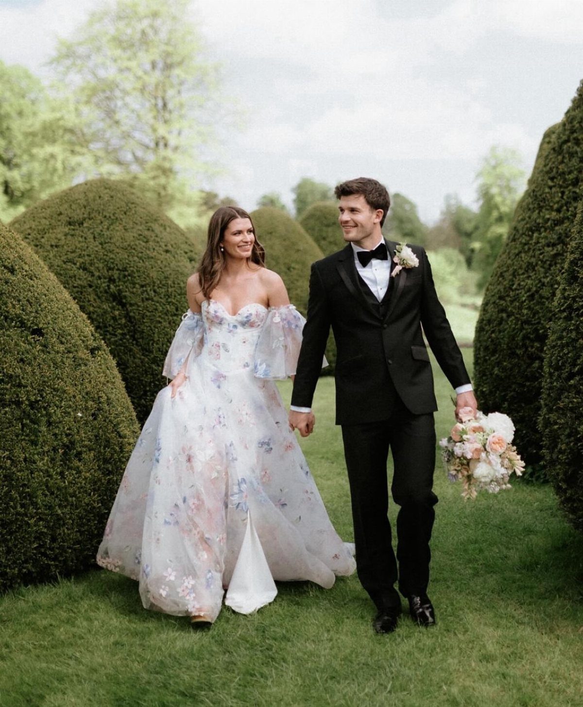 A bride in a floral patterned wedding dress and a groom in a tuxedo walk through a garden.