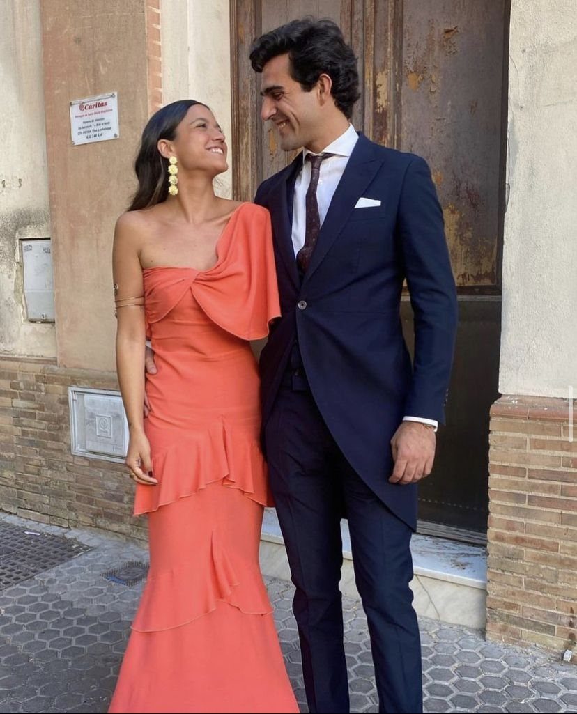 A man in a navy suit and a woman in a coral gown smiling at each other.