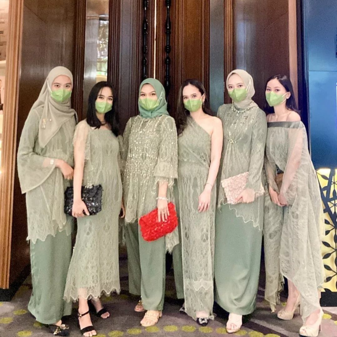 Six women posing in matching sage green lace dresses and hijabs wearing green face masks.