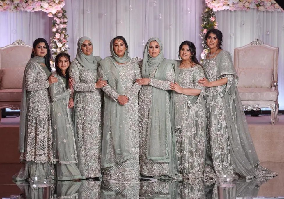 Seven women posing in matching sage green embellished traditional South Asian formal dresses and hijabs.