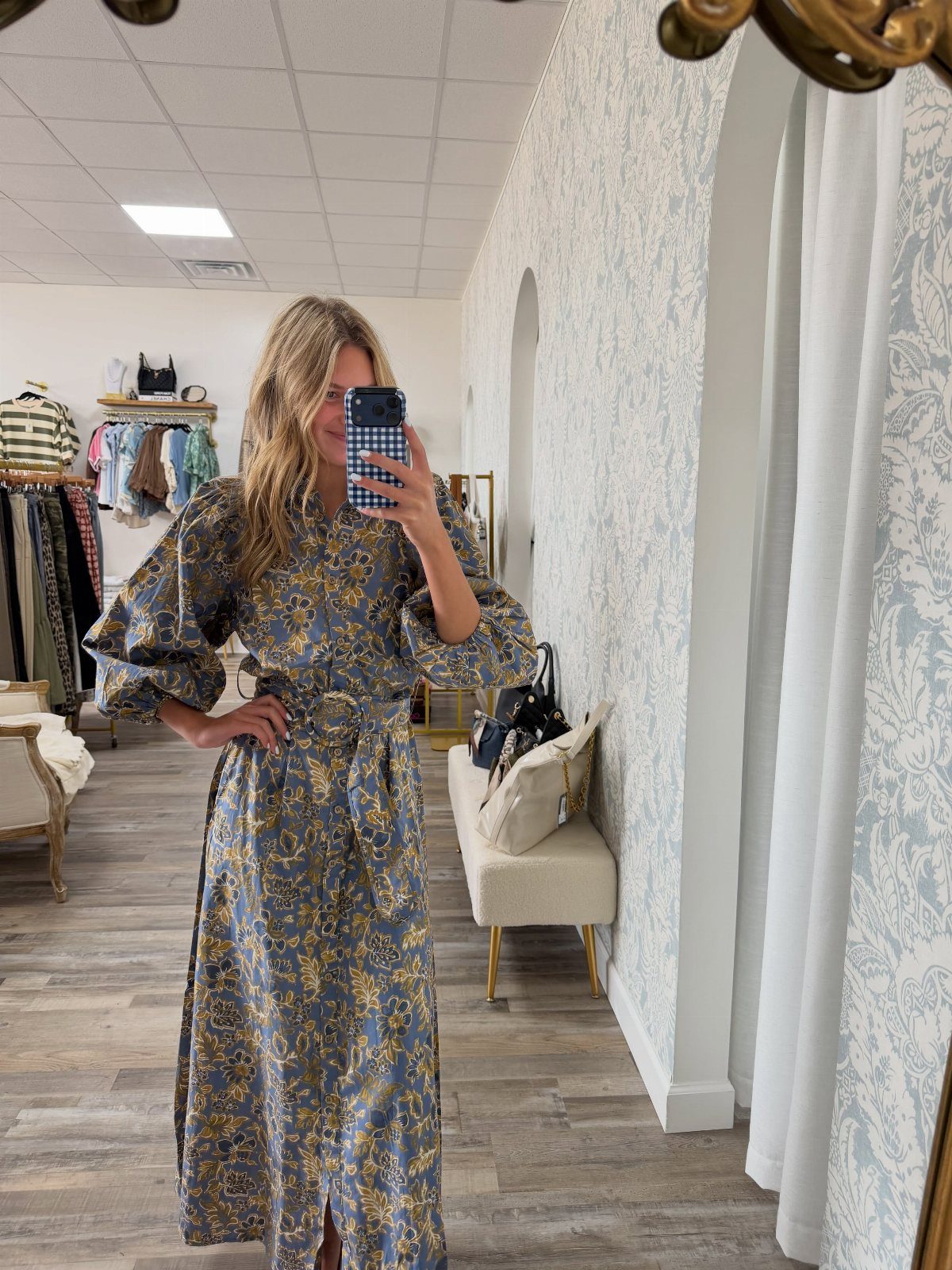 Woman in a blue and gold patterned maxi dress taking a mirror selfie in a boutique.
